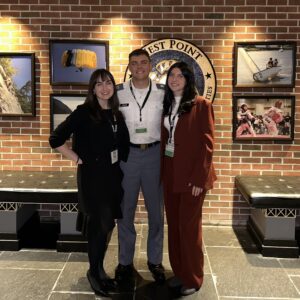 Two female college students stand on either side of a West Point cadette. They are inside, and a brick wall is behind them.