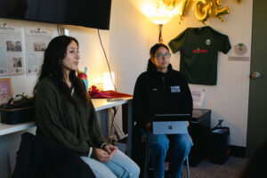 Two college students sit in an office. One holds a laptop on her lap.