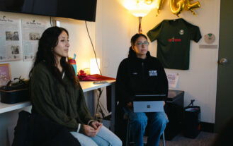 Two college students sit in an office. One holds a laptop on her lap.