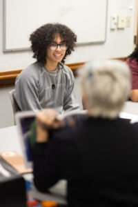 A male college student sits at a table and listens to someone standing in front of him.