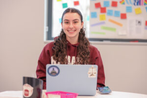 A student wearing a maroon Ramapo sweatshirt sits at a table. A laptop adorned with stickers is open in from of her. She is smiling at the camera.