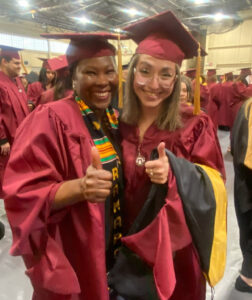 Two women stand side by side , smiling, in maroon graduation gowns and caps.
