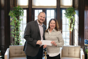 A man and a woman stand together in front of a bright window. They are holding a check.