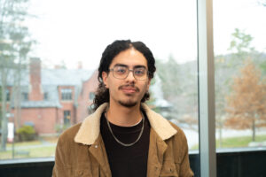 A male college student with glasses stands in front of a window.