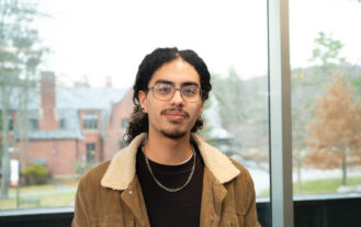 A male college student with glasses stands in front of a window.