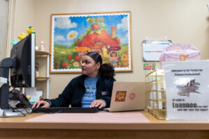 A young woman sits behind a large desk, with a laptop to one side, and a desktop to the other. She uses the keyboard and looks at the monitor. A large colorful picure is on the wall behind her.
