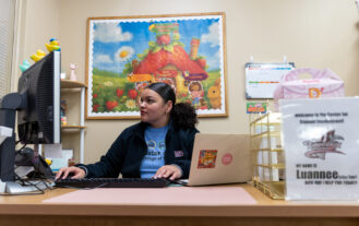 A young woman sits behind a large desk, with a laptop to one side, and a desktop to the other. She uses the keyboard and looks at the monitor. A large colorful picure is on the wall behind her.