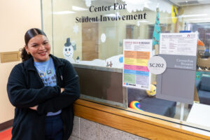 A young woman stands smiling with her arms crossed. She is in front of a window that states Center for Student Involvement.