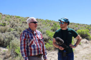 A man in headphones, holding a microphone, inerviews stands and interviews an older man outside.
