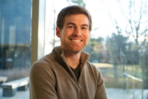 A man in a brown sweater smiles in front of a window.