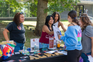 Three young women stood behind a table outside, as another woman stood infront of it and speaks with them.