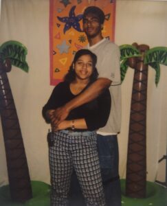 A man and a woman stand together, with the man's arm around the woman. They are flanked by two blowup palm trees.