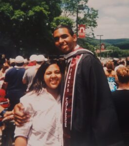 A man and a woman stand closely together. The man, dressed in a black graduation gown and maroon stole, has his arm around the woman. They are outside in front of trees.