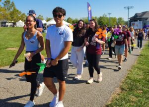 A group of people walk together on a sunny day along a park path, holding orange pinwheels and wearing casual clothes, as part of an organized outdoor event.