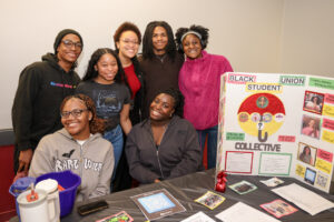 Six smiling students pose together behind a table with informational materials and a colorful poster for the Black Student Union. The table has flyers, a sign-up sheet, a QR code, and a blue pitcher.