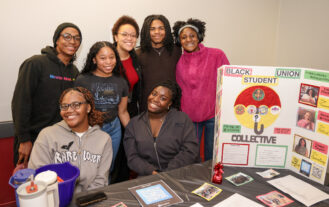 Six smiling students pose together behind a table with informational materials and a colorful poster for the Black Student Union. The table has flyers, a sign-up sheet, a QR code, and a blue pitcher.