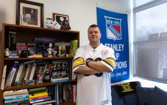 A man in a football jersey stands with his arms crossed in front of a bookcase.