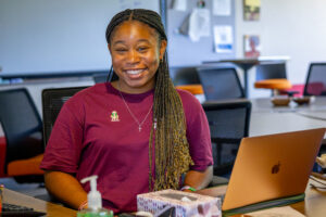 A woman in a maroon T-shirt sits at at a table with a laptop open. She is smiling.