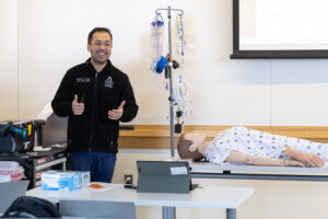 A man stands in a classroom giving thumbs up beside a medical mannequin on a table, with medical equipment and a projector screen in the background.
