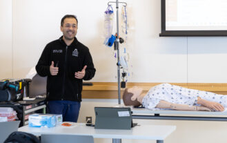 A man stands in a classroom giving thumbs up beside a medical mannequin on a table, with medical equipment and a projector screen in the background.