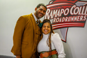 A man and a woman stand closely togehter, smiling, in front of a sign that reads Ramapo College.
