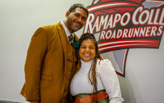 A man and a woman stand closely togehter, smiling, in front of a sign that reads Ramapo College.