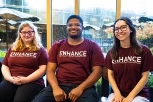 The college students in maroon t-shirts with the word ENHANCE on them sit next to eachother in front of a large window.