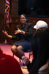 A woman sitting on a chair wearing a black suit speaks. A college student in a white hat sits in front of her.