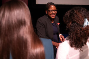 A woman in a black suit smiles as she introduces herself. She is flanked between the back of two heads of brown hair.