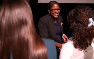A woman in a black suit smiles as she introduces herself. She is flanked between the back of two heads of brown hair.