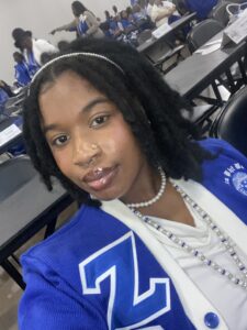 A person with short dreadlocks, wearing a blue and white jacket with a large Z, a pearl necklace, and a nose ring, takes a selfie at an indoor event with others in similar blue attire seated behind them.