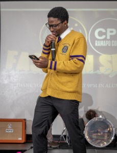 A young man wearing glasses and a yellow cardigan with purple stripes reads from his phone while speaking into a microphone on stage. Musical equipment and a drum set are visible in the background.