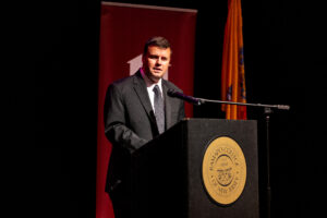A man in a suit stands at a podium on stage. A maroon banner is behind him.