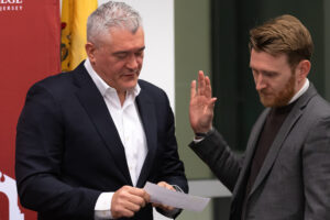 BOT Chair Albert Nieves stands to the left of Trustee O'Connor, pointing to the words O'Connor, who stands holding his right hand in the air, needs to recite as he is sworn in to his role as a trustee.