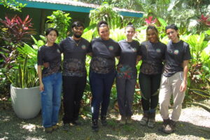 Six people stand side by side, smiling and wearing matching black shirts with colorful patterns, in a lush garden area with green plants and trees surrounding a building in the background.
