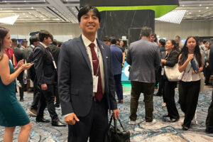 A man in a dark suit stands smiling with a bag in hand at a busy conference event. People are mingling in the background.