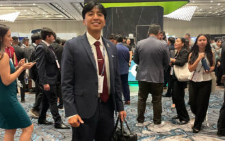 A man in a dark suit stands smiling with a bag in hand at a busy conference event. People are mingling in the background.