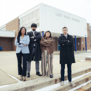 Four adults stand confidently with arms crossed outside a building labeled Lodi High School, on a cloudy day. They are dressed in business and casual attire, facing the camera and standing on concrete steps.