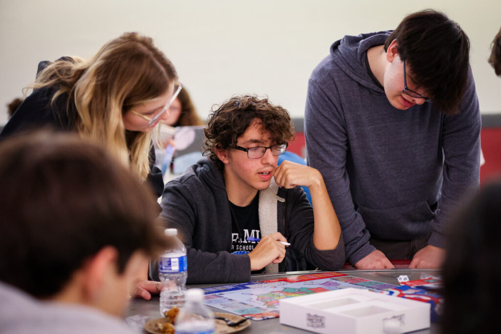 A group of teenagers gathers around a table, focused on playing the board game Election Night. One holds a pencil, others lean in with interest. A water bottle and food are on the table, and the atmosphere appears collaborative and engaged. They gathered together on Election Night, November 5, 2024, to monitor live updates on national and local elections.