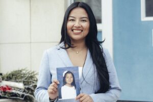 A smiling woman with long dark hair stands outside, holding a photo of a young girl wearing a white outfit and hair bow. The background features a blue door and part of a building.