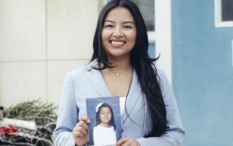 A smiling woman with long dark hair stands outside, holding a photo of a young girl wearing a white outfit and hair bow. The background features a blue door and part of a building.