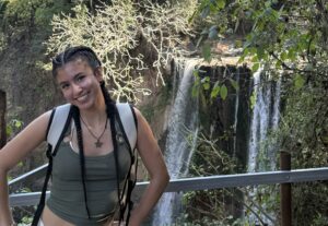 A smiling woman with braided hair, wearing a green crop top and a backpack, stands on a railing in front of a lush forest and a tall waterfall cascading down the rocks in the background.
