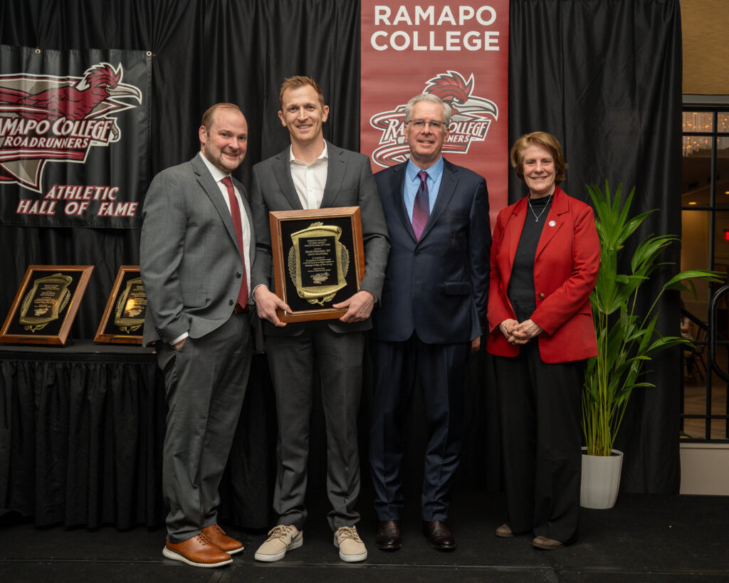 Shane Donohue stands with Vice President Chris Romano and President Cindy Jebb in front of a black curtain with Ramapo College Athletic Hall of Fame signage. One person holds a plaque. They are dressed in formal attire and smiling at the camera. A plant is on the right.