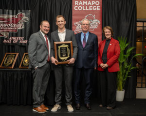 Shane Donohue stands with Vice President Chris Romano and President Cindy Jebb in front of a black curtain with Ramapo College Athletic Hall of Fame signage. One person holds a plaque. They are dressed in formal attire and smiling at the camera. A plant is on the right.