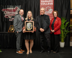 Michelle (Micklos) Hines stands on a stage at a Ramapo College event. The woman second from left holds a plaque. Banners and plants are in the background. All are dressed formally and smiling at the camera.