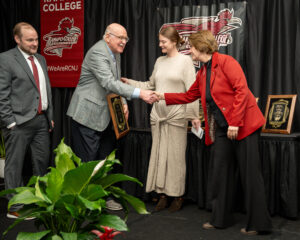 Four people stand on a stage at a Ramapo College event. Two are shaking hands while holding plaques, with two others, a man and a woman, standing nearby. A plant is in the foreground and college banners hang behind them.