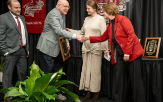 Four people stand on a stage at a Ramapo College event. Two are shaking hands while holding plaques, with two others, a man and a woman, standing nearby. A plant is in the foreground and college banners hang behind them.