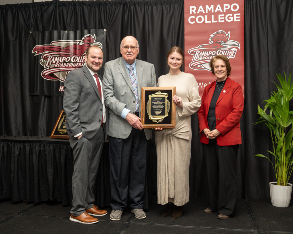 Lindsay (Hughes) Neumann stands indoors in front of Ramapo College banners with her grandfather, VP of Outreach & Engagement Chris Romano, and President Cindy Jebb. Lindsay and her grandfather hold a large plaque. All are dressed formally and smiling at the camera. A potted plant is visible on the right.