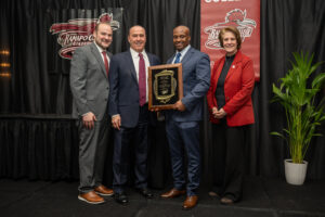 Antione Pryor stands on a stage; one man holds a large plaque. Ramapo College banners are displayed behind them, and a potted plant is on the right side of the stage.