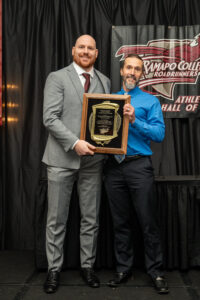 Craig Van Leeuwen and his coach stand on stage, smiling and holding a large plaque. One wears a grey suit and the other a blue shirt. They are on a stage with a black curtain and a Ramapo College Roadrunners banner behind them.